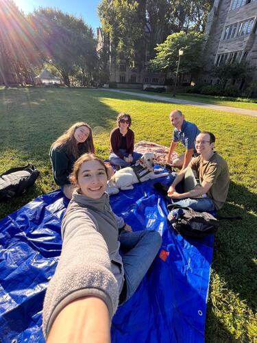 three students sitting on the grass with professor sarah glenski and Yale's therapy dog, Heidi, along with Heidi's handler